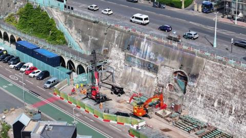 A large section of concrete wall as seen from the air with blue railings and some of the blue archway pieces laid to one side next to bright orange construction equipment on Brighton promenade