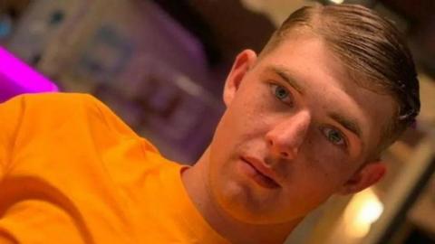 A man in an orange t-shirt looking at the camera from a close distance. He is white and has short blonde hair. His hair is slicked to the right. The background is blurred with pink lighting to the left.