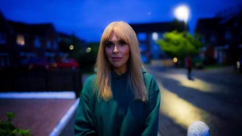 A woman with long, blonde hair and a green jumper outside in the street at dusk.
