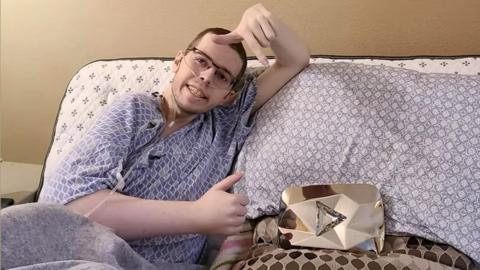 A young man lies on his side in a hospital bed, one thumb up and the other above his head, pointing downwards. He's wearing a hospital gown and has an oxygen tube in his nose. Next to him on the bed is a gold helmet with a silver triangle stuck to the front.