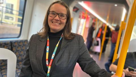 Nexus managing director Cathy Massarella smiling as she hold a yellow hand rail while on board the first new Tyne and Wear Metro in December 2024. She is wearing grey suit jacket over a black turtle neck top. She has glasses and a colorful lanyard. Her brown hair curls at the ends  and reaches her shoulders. Behind her two men are sitting and looking in their phones. Other passengers are standing further back in the train. 