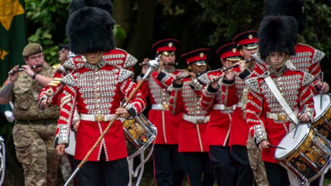 A military parade with soldiers in red ceremonial uniform with metal buttons and large fur hats or caps. They are holding or playing drums or wind instruments. 