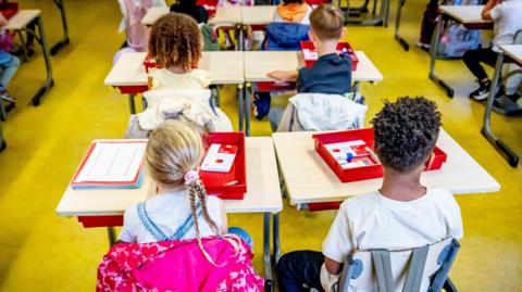 Young children are sitting at wood-topped desks in a classroom and they have their backs to the camera. Their coats are hung over the backs of their chairs and on their desks are plastic boxes which have pads of paper and coloured pens. 