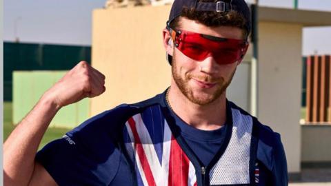 Denzil Grose is smiling at the camera and raising his right hand in a victorious fist. He is wearing red sunglasses and a GB team waistcoat which features a Union Jack. He has dark hair and a beard. Behind is part of the shooting range.