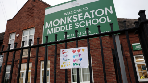 An exterior view of a red brick school with black railings outside. A large green sign with white text reads 'Monkseaton Middle School' and a hand-drawn picture attached to the railings reads 'Save this school'. 