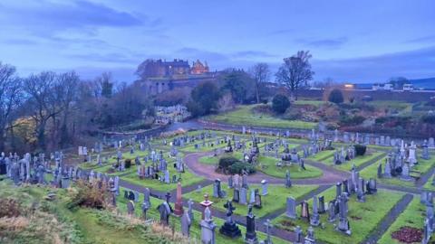 A mid-blue dusky sky with a few darker blue clouds, sits over a graveyard with a castle on the horizon.