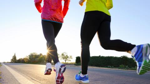 Two people running on a road in bright jackets, leggings and trainers. Shot from the waist down. 