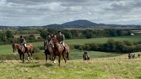 In the foreground are three horses being ridden by hunters wearing tweed jackets, beige jodhpurs, and long black riding boots. The horses are various shades of brown and chestnut - there are more riders and horses in the background. They're riding along a ridge with the Wrekin clearly seen in the background - the weather is sunny and the grass is bright green. 