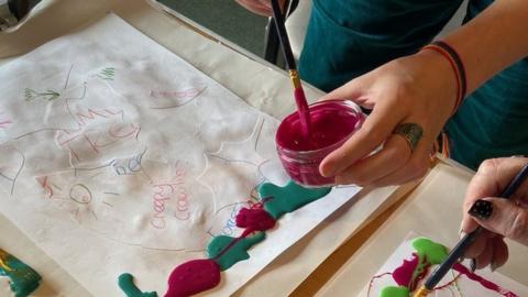 A piece of artwork with a person's hands in shot, dipping a paintbrush into pink paint