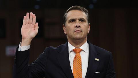 A man in a dark suit and orange tie raises his right hand as he looks forward.
