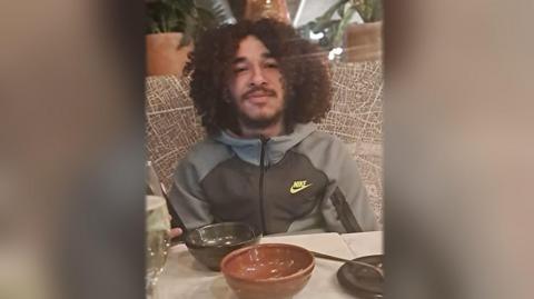 An image of a man with dark, curly, shoulder-length hair. He is wearing a grey Nike zip top and is sat at a table with some empty bowls on. He is looking at the camera.