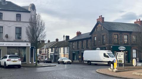 A street view of Kilrea.  There are shops in the background and traffic is approaching a roundabout in the foreground.  Cars are also parked outside shops. 