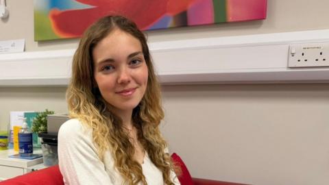Molly looks directly into the camera. She has curly blonde hair and dark blue eyes and is wearing a white blouse.