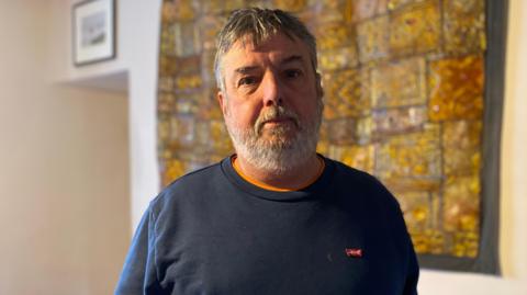 Peter Roffey stands indoors wearing a navy sweatshirt with a small logo, positioned before a warm-toned patterned artwork. A sofa, lamp, and doorway appear in a softly lit, blurred background.