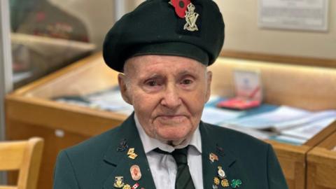 Albert, sitting present day, wearing a green beret with Royal Ulster Rifles insignia. He is wearing a green uniform with a number of poppy and military pin badges 