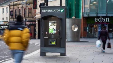 A high street showing a row of shops and a communication hub in the middle of the pavement. It looks like a dark grey phone box and says "defibrillator" on the top. The blurry figures of two people are walking past