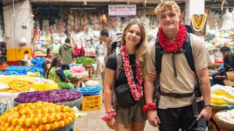 Contestants Sioned and Fin are in a busy and colourful market Indian market. They are both wearing backpacks.