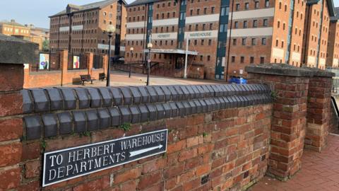 Herbert, Kimberley and Philpott warehouses at Gloucester Docks. It is a red brick building with a short brick wall in the foreground.