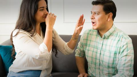 A woman holding her nose and putting out her hand in front of her face while a man talks