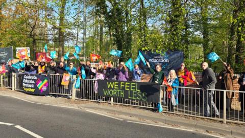 Dozens of people waving blue flags stand at the side of a road in a protest. There are banners reading 'we feel worth less' and advertising the NEU.