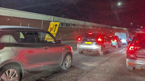 Two lanes of cars queue on a road. A yellow sign sits to the right of the road that shows one arrow pointing ahead, and another next to it erased out.