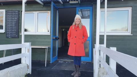 Lynda stands in front of the pavilion smiling wearing a red winter coat, brown boots and woolly hat