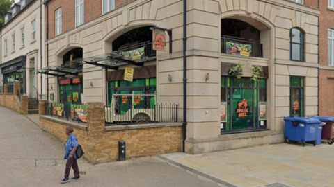 The outside of a restaurant called Jurassic Grill. A woman is walking past. There is a large blue bin on the right and green signs in the windows. There is a wall with railings in front of the building.