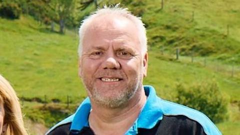 Mark Williams stands in a field. Mark has close-cropped grey hair and a short beard and he's smiling into the camera. He is wearing black and blue T-shirt.