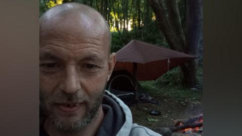 Blair Murray has a shaved head with a stubbly beard. He is wearing a grey hoodie and stands in front of a fire and tent. His tent has a tarpaulin strung over his tent in the middle of woodland. The picture is a selfie.