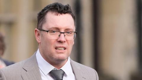 Image shows man in a light-brown suit with a silver tie and black-rimmed glasses. He has black hair and grey stuble.