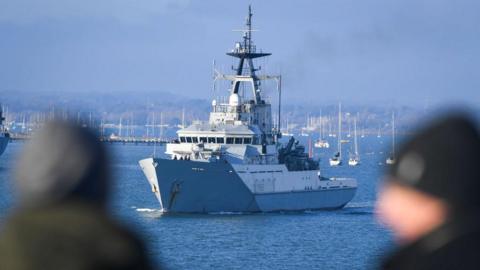 HMS Tyne leaves Portsmouth on December 31, 2020. The naval vessell is in the midground of the image with two people's heads out of focus looking towards the ship in the foreground. The sky and sea are blue on a clear day and there are moored yachts and yachts on a pontoon in the background