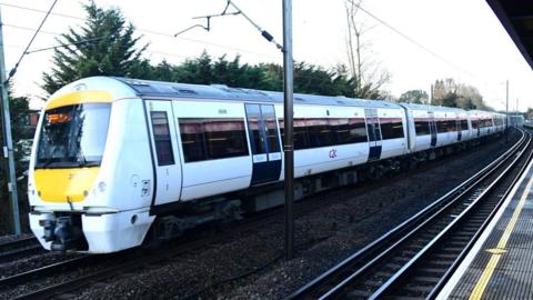 A c2c train, which is white with a yellow front, passes through a station on a cloudy day.