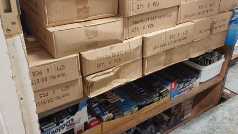 Lots of cardboard boxes stacked in a backroom. On the bottom shelves are packets of cigarettes and vapes. 