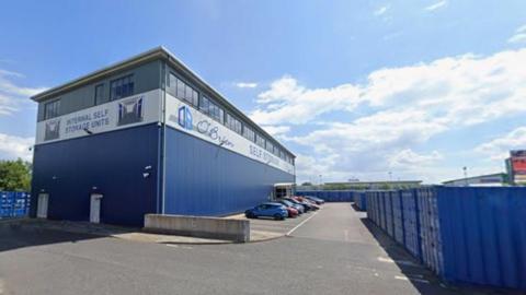 An existing storage yard owned by O'Brien on Ferryboat Lane. A large blue industrial office building stands to the left. There is a row of cars parked outside. Around three sides of the building are rows of blue storage containers.