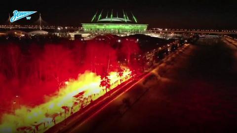 A road looks like it is on fire from flares leading to a green-lit football stadium