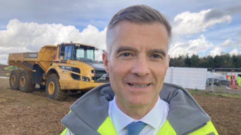 Councillor Mark Harper standing by a digger and wearing a high vis jacket