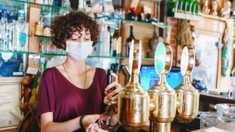 A young woman with curly hair working in a bar pulling a pint of beer from one of the taps while wearing a face covering