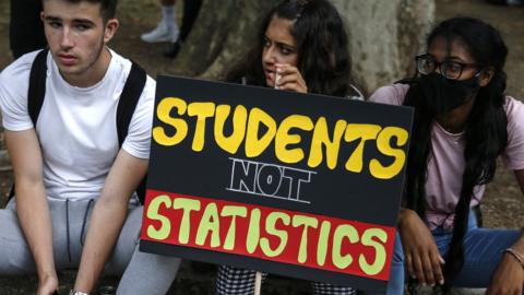 Three students sit on a street kerb with a protest placard reading: "students not statistics"