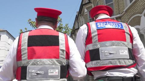 Two Bobbies, wearing red vests, red hats and white shirts, patrol on St John's Road in London.