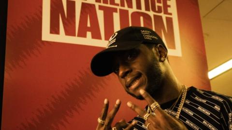 The rapper 6lack stands in front of a signt that says 'Apprentice Nation'
