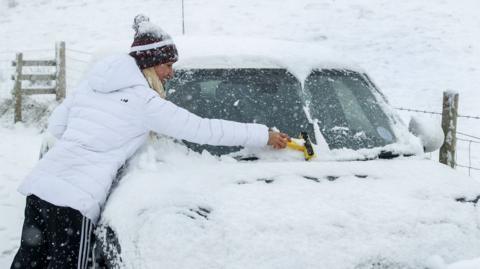 A woman uses a scraper to clear her windscreen amid heavy snow