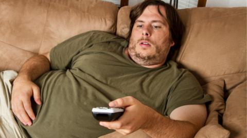Overweight man with a remote control in his hand slouched on a sofa