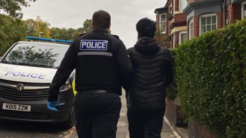 A suspect in black clothes is led to a police van by an officer