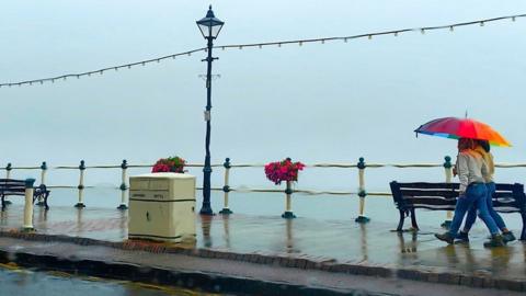 Two women walk along the sea front holding and open rainbow-coloured umbrella over them. The sea and sky behind are merged together in shades of pale blue.