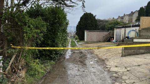 Police cordon in place down Dransfield Way in Bath. Hedges and houses can be seen either side of the path.