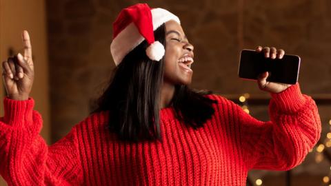A woman wearing a Santa hat mimes singing along to music, using her phone as if it was a karaoke microphone