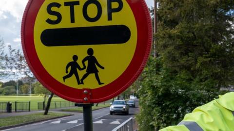 The arm of a crossing guard, known as a lollipop man or lady in the UK, preparing to stop traffic so school children can cross the road safely.