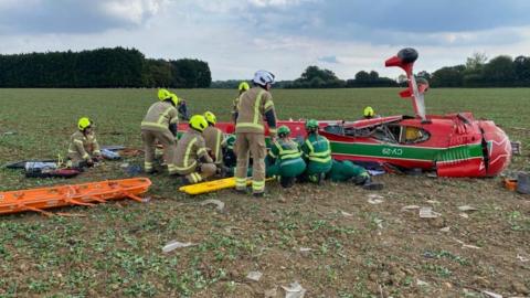 A red light aircraft upside down. Wheels are sticking up and it has been surrounded by emergency services including paramedics and firefighters