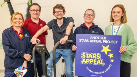 Charles McGrath on a cycling machine surrounded by charity representatives