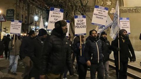 Group of drivers wearing winter clothes and carrying placards saying e.g. "stop racist policies"